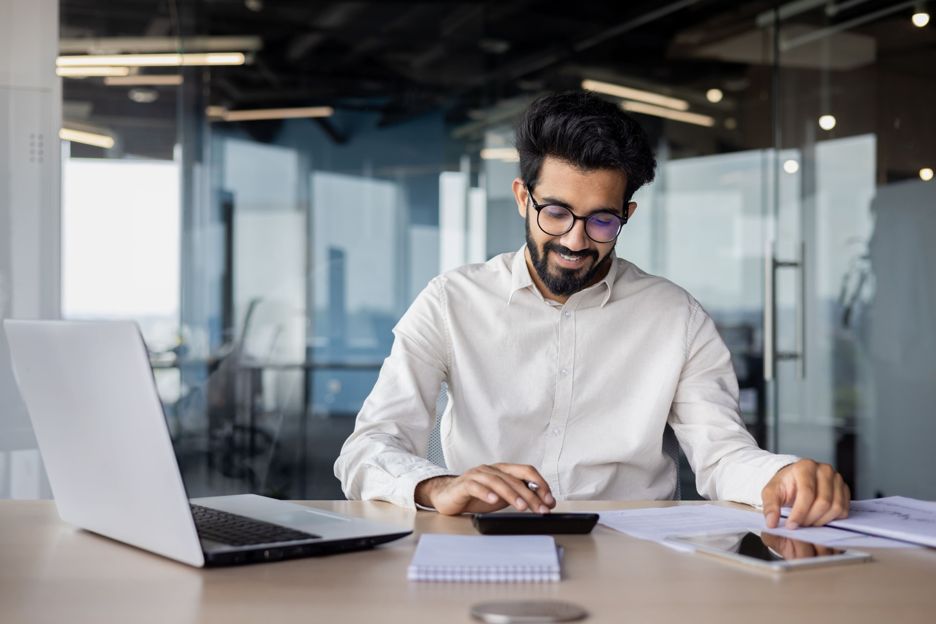 Indian young male accountant, financial analyst and auditor working focused in office at desk with documents and calculator